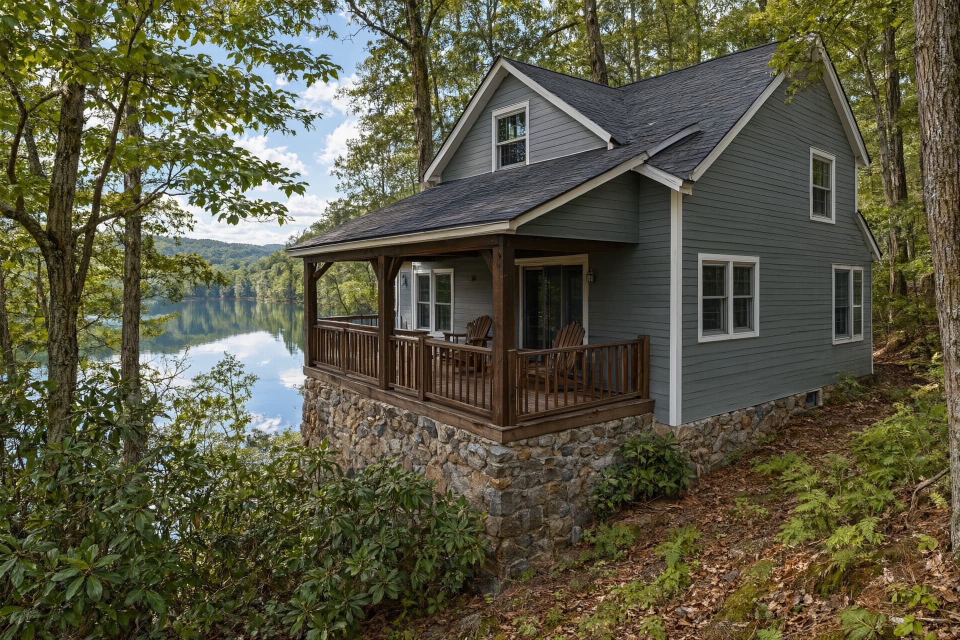Gray two-story lake house with a covered wood porch on a wooded slope above a lake