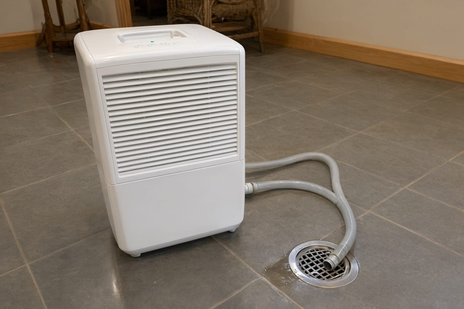 White portable dehumidifier on a tile floor with its drain hose running into a floor drain