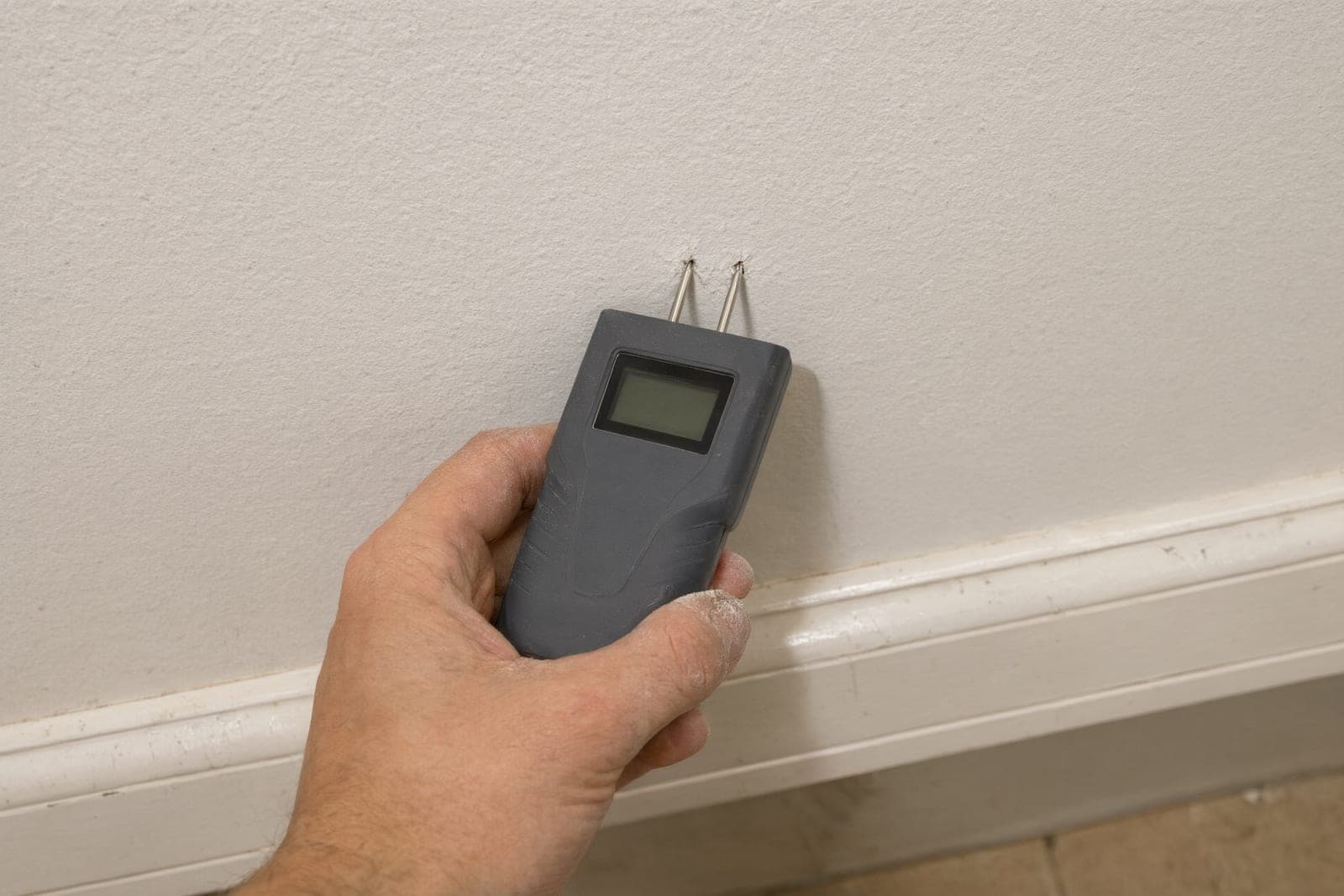 Hand pressing a plain handheld moisture meter into drywall just above the baseboard